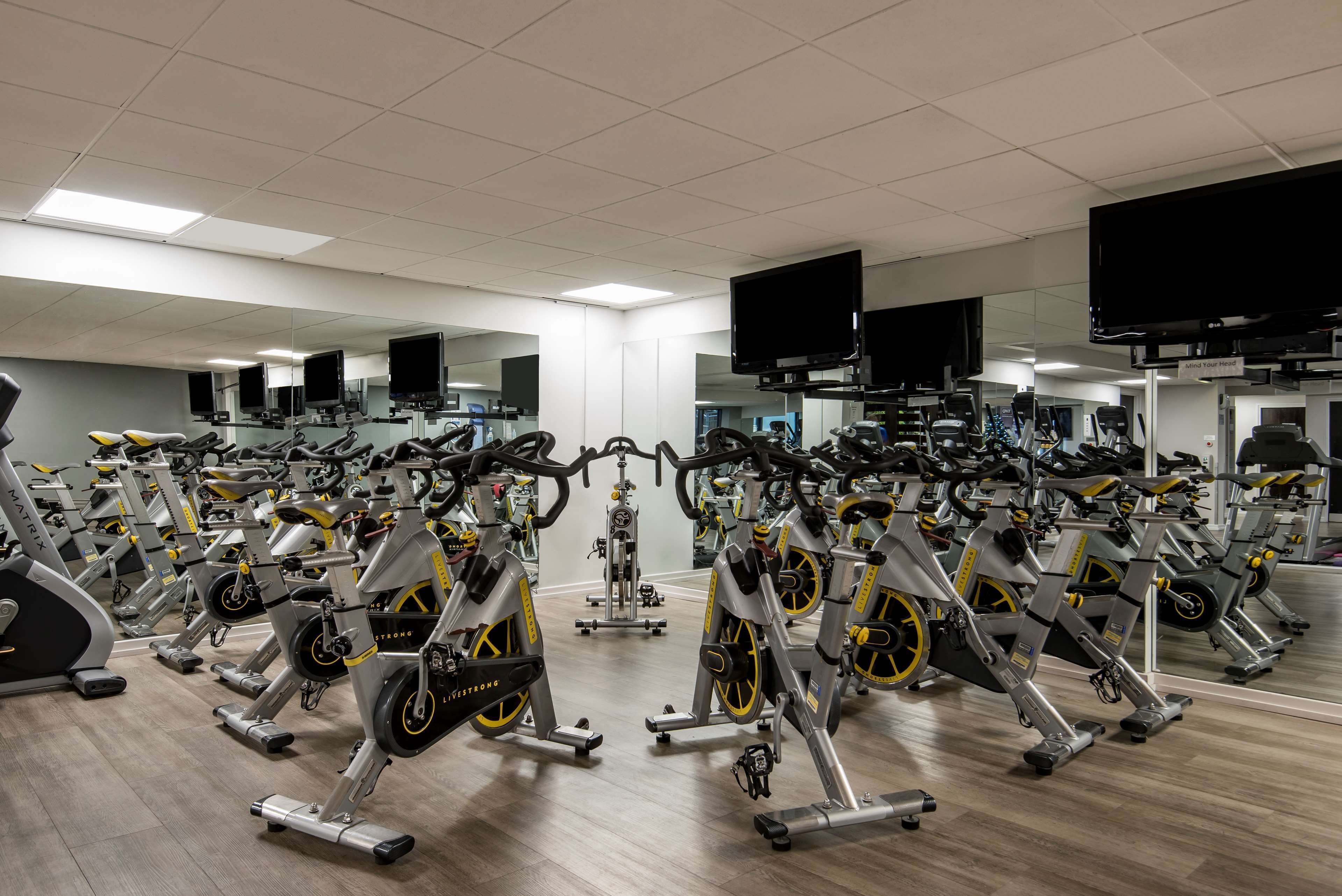 A gym with multiple stationary bikes arranged in rows, large mirrors on the walls, and ceiling-mounted televisions.