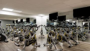 A gym with multiple stationary bikes arranged in rows, large mirrors on the walls, and ceiling-mounted televisions.