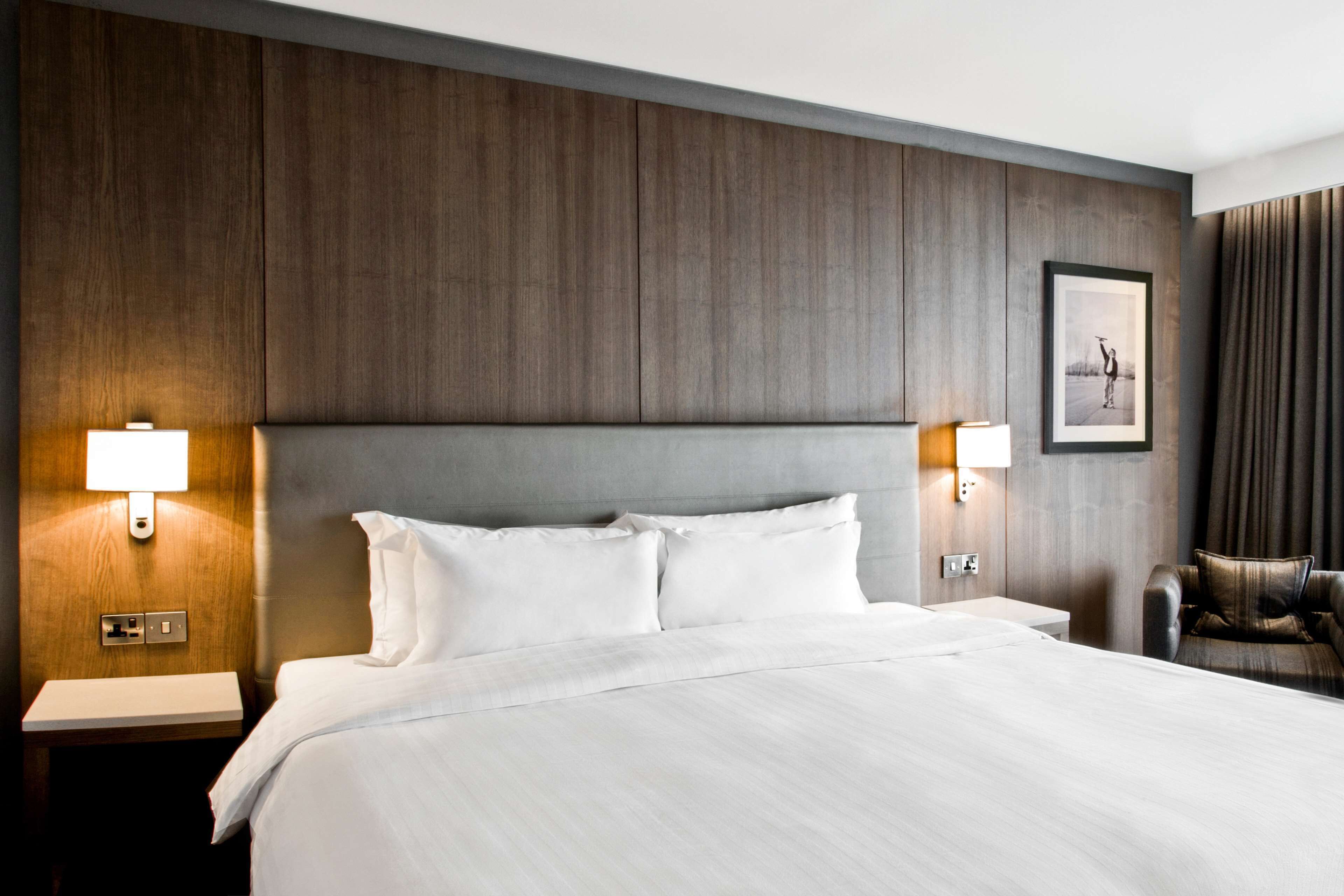 Una habitación de hotel con una cama grande, dos mesitas de noche, una silla y un cuadro en la pared.
