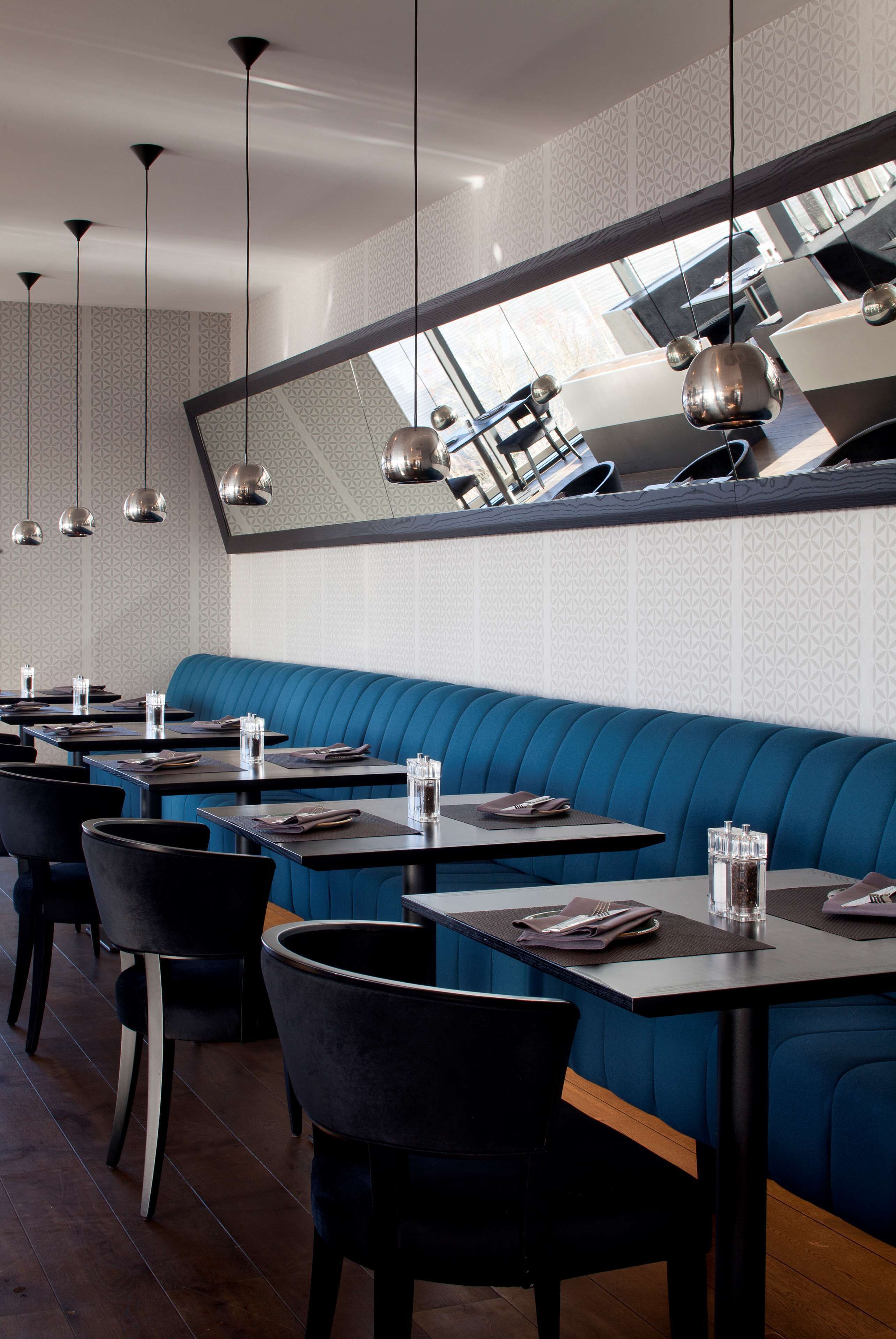 A restaurant interior with a long blue booth seating area, multiple tables with chairs, hanging pendant lights, and a patterned wall.