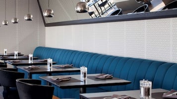A restaurant interior with a long blue booth seating area, multiple tables with chairs, hanging pendant lights, and a patterned wall.