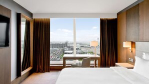 A hotel room with a large bed, a desk, and a view of a cityscape through a large window.