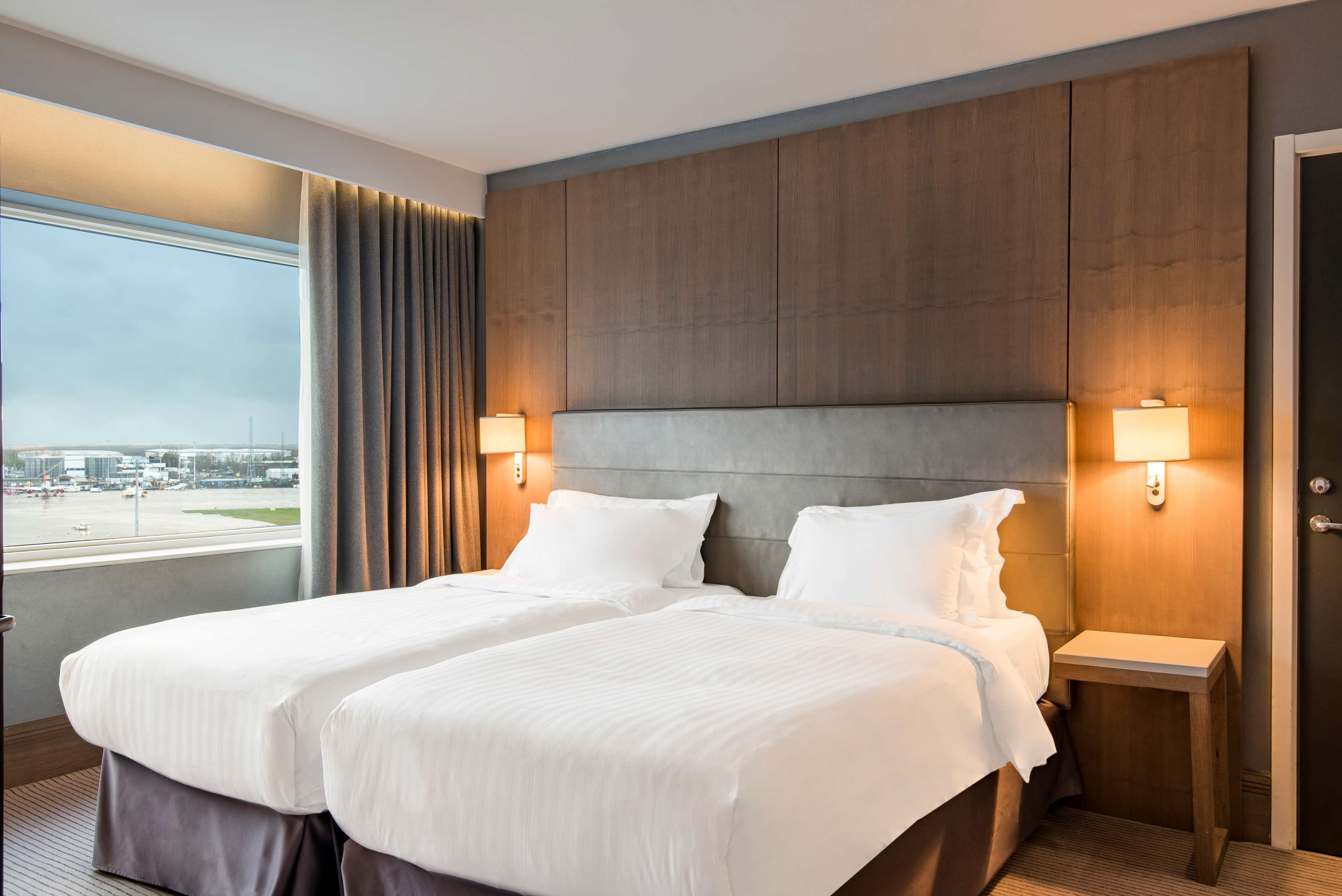Une chambre d’hôtel avec deux lits, une grande fenêtre donnant sur un aéroport et des panneaux en bois au niveau de la tête de lit.