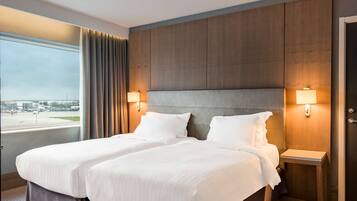 Une chambre d’hôtel avec deux lits, une grande fenêtre donnant sur un aéroport et des panneaux en bois au niveau de la tête de lit.