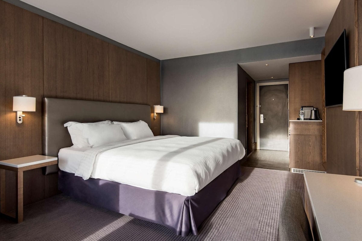 A modern hotel room with a large bed, bedside tables, a flat-screen TV, and a kitchenette area.