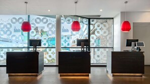 A reception desk area with two dark-colored desks, each with a monitor and a red pendant light. The area is well-lit with natural light coming through large windows with a circular pattern design.
