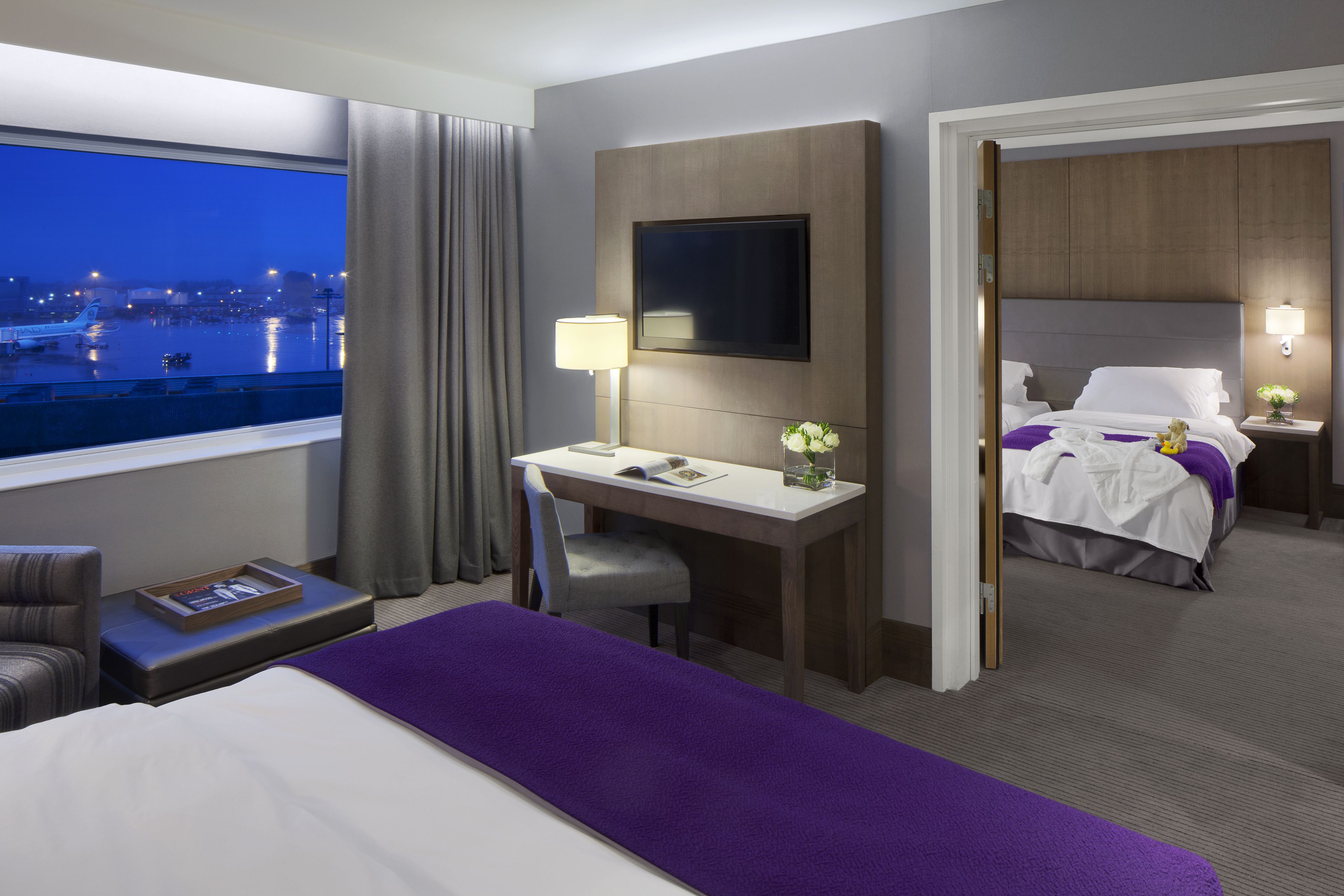 A hotel room with a desk, a bed, a TV, and a view of the airport.