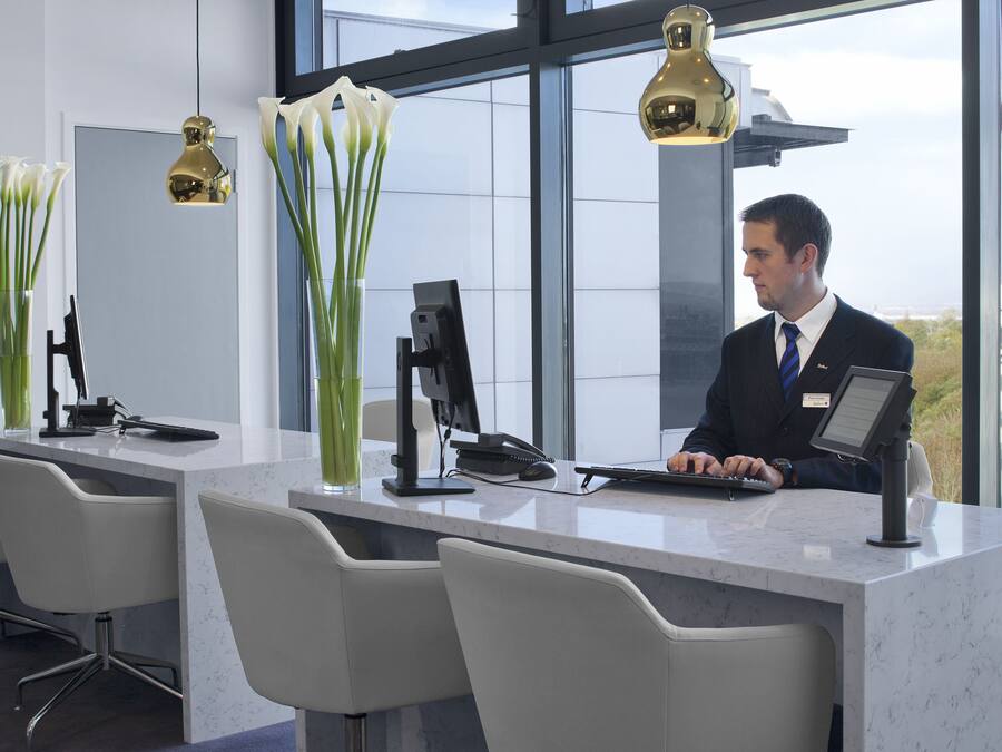 A modern reception desk with a computer, a phone, and a cash register. There are three chairs and two vases with flowers.