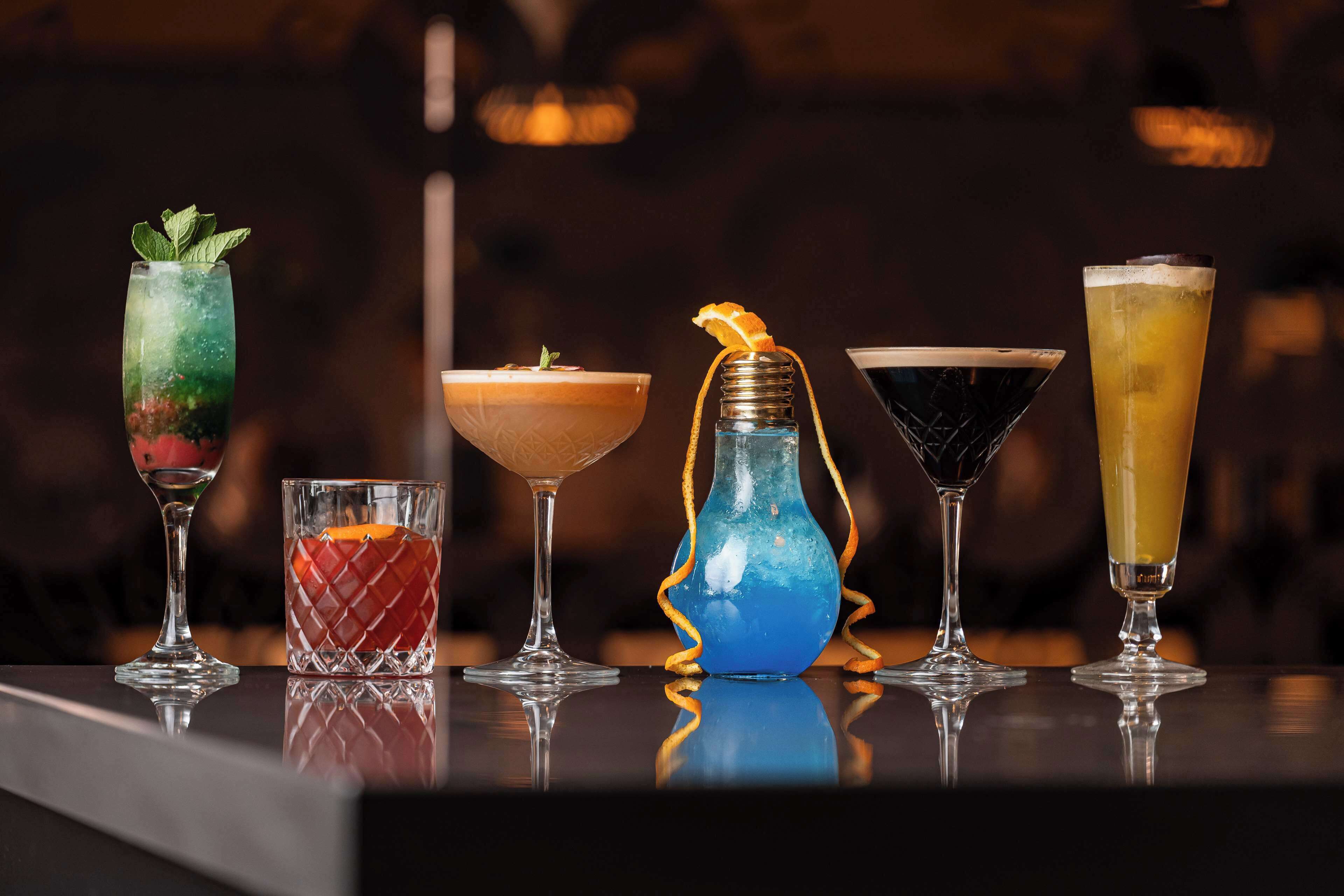 A bar with various drinks, including a cocktail with a mint garnish, a blue drink in a vase, and a tall glass with a yellow drink.