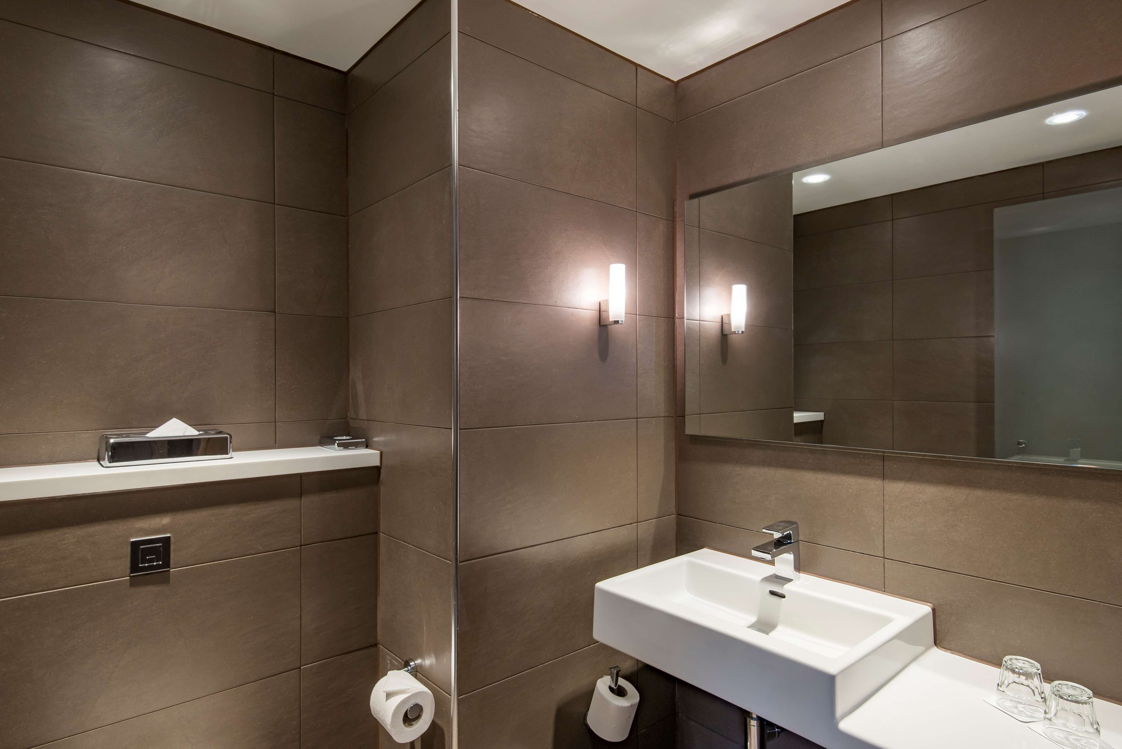 A modern bathroom with a large mirror, a white sink, and a toilet paper holder.