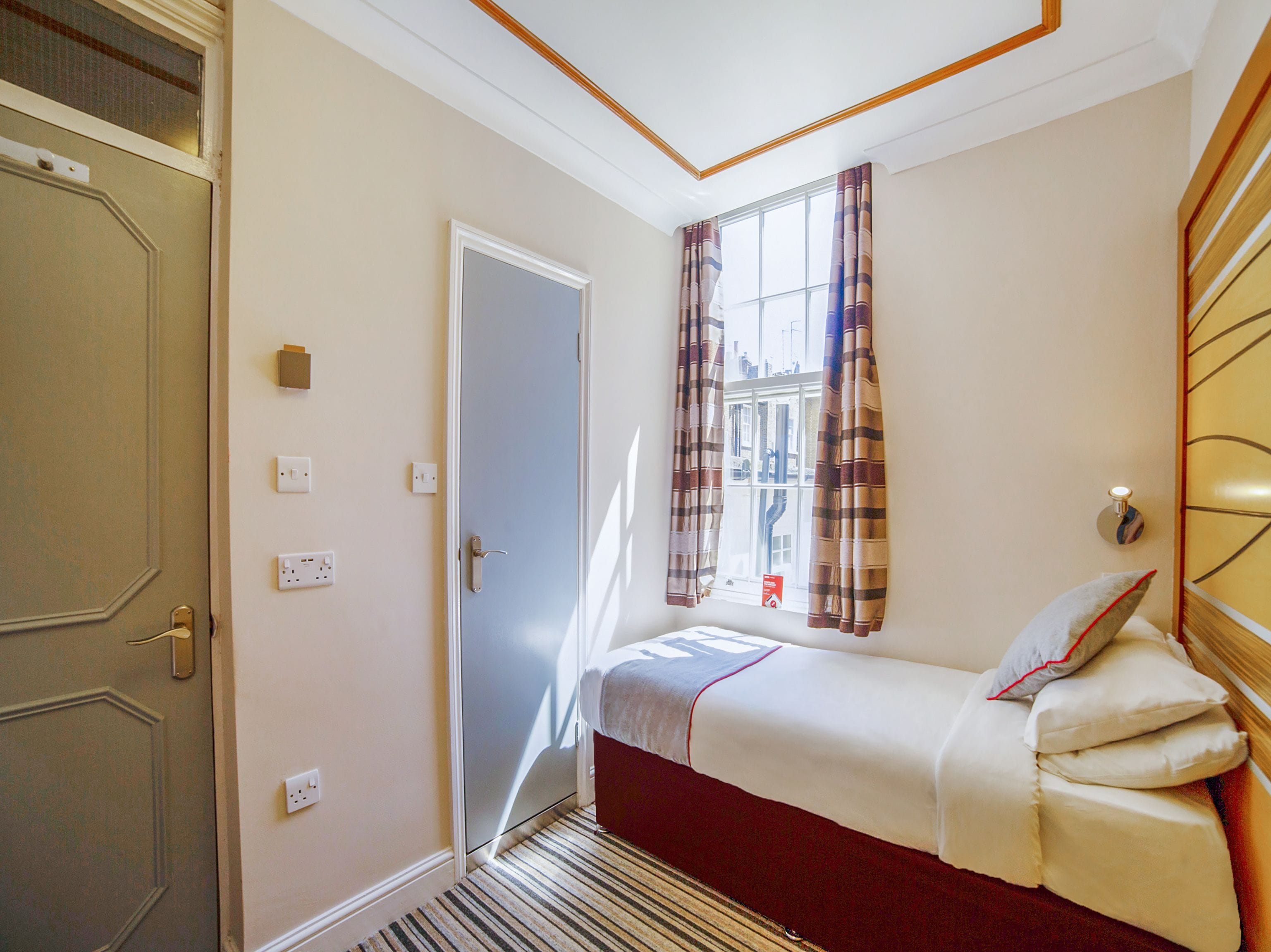 A hotel room with a single bed, a window with curtains, a door, and a wall-mounted light fixture.