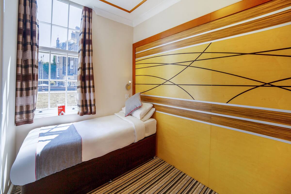 A hotel room with a bed, a window with curtains, a wall with a geometric design, and a striped carpet.