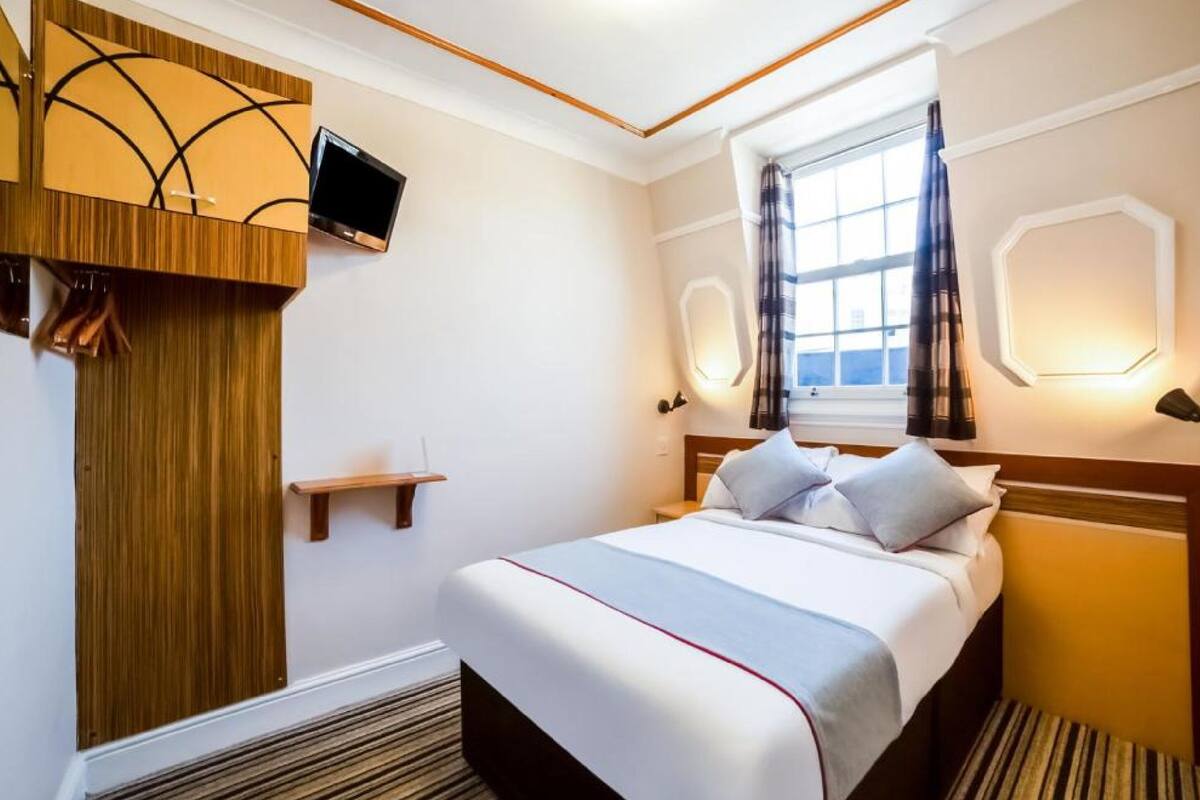 A hotel room with a bed, a TV mounted on the wall, a window with curtains, and a wooden panel on the wall.