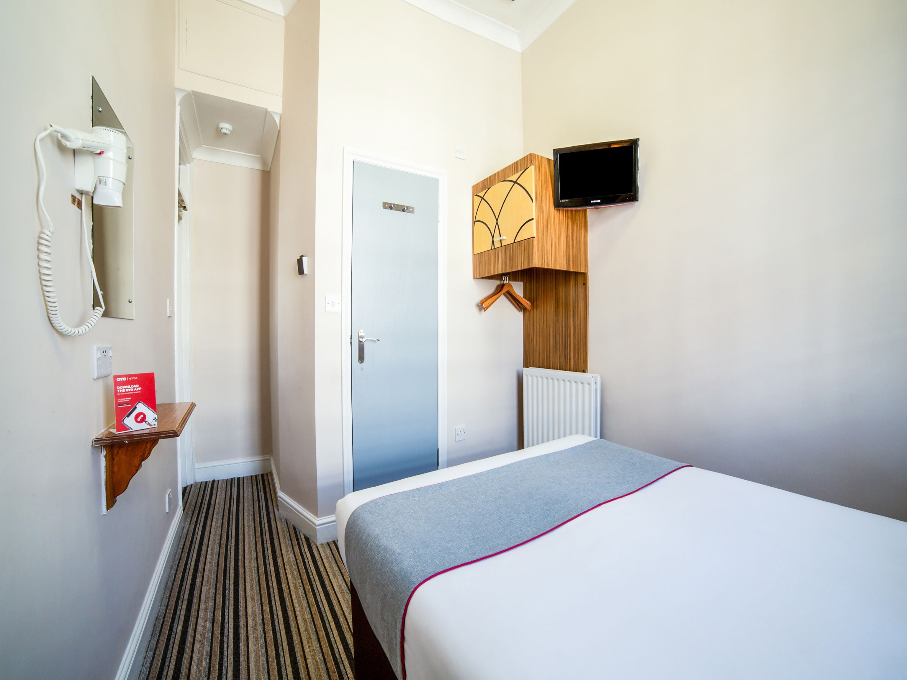 A hotel room with a bed, a TV mounted on the wall, a radiator, a coat hanging on a wooden rack, and a hairdryer on the wall.