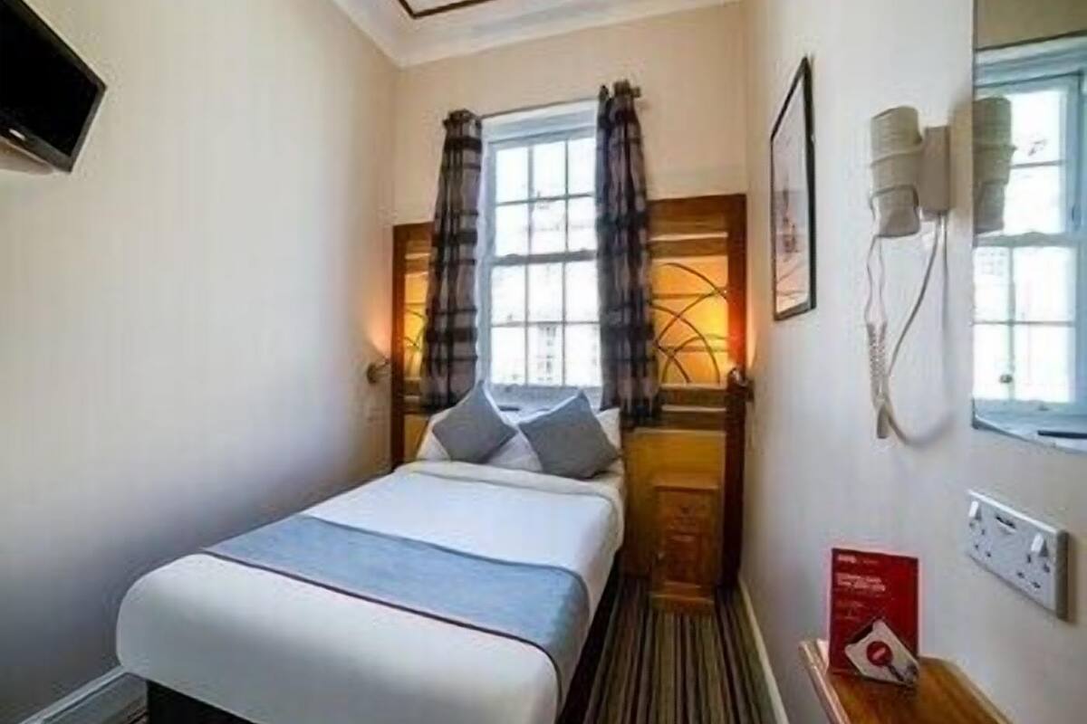 A hotel room with a bed, a window with curtains, a bedside table, a wall-mounted phone, and a light fixture.