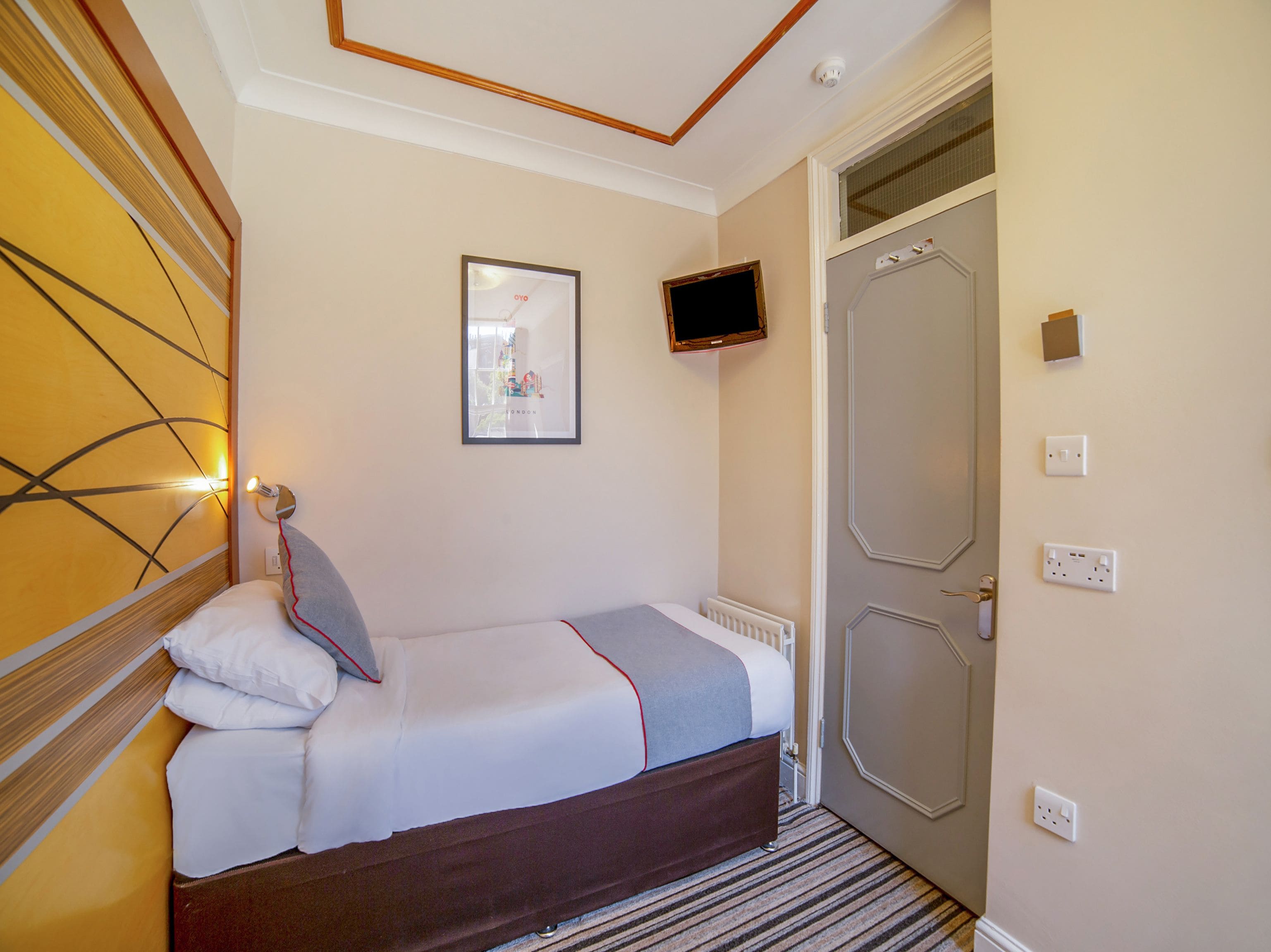 Habitación de hotel con una cama, un cuadro enmarcado en la pared, un televisor sobre la cama y una puerta con manija.