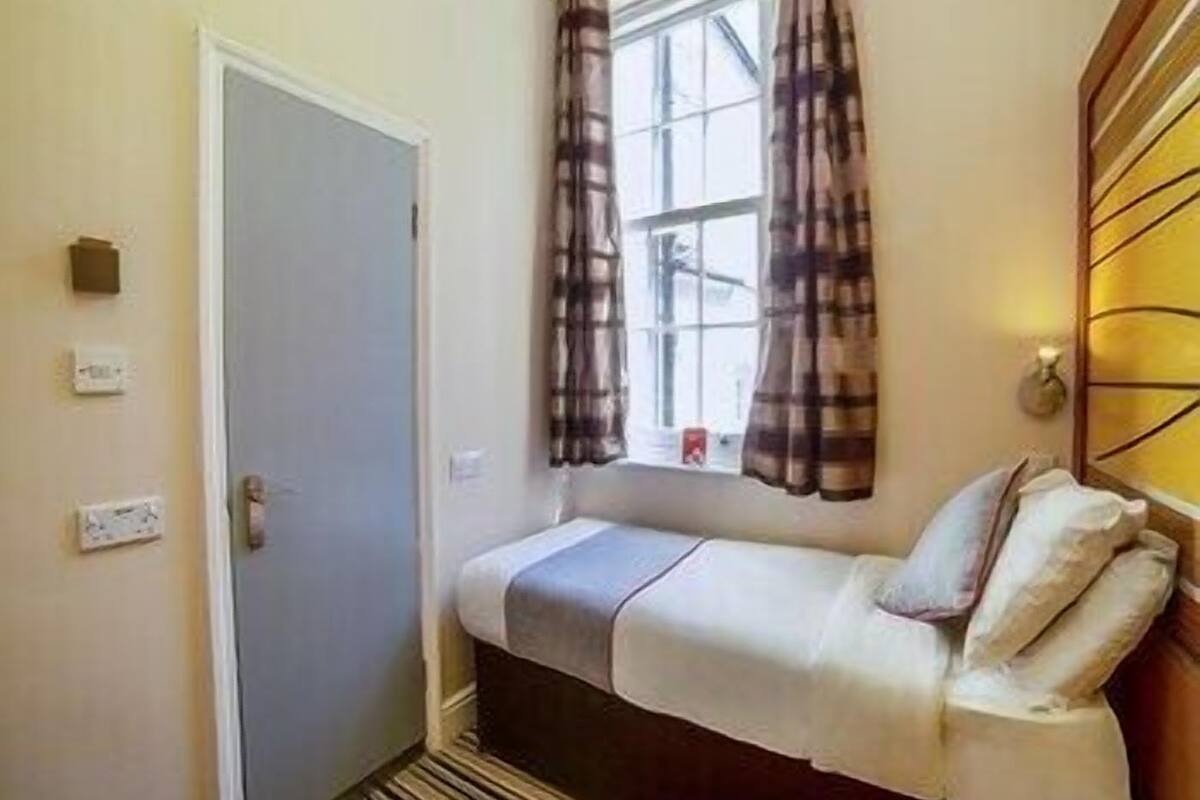 A small room with a bed, a window with curtains, a door, and a wall-mounted light fixture.