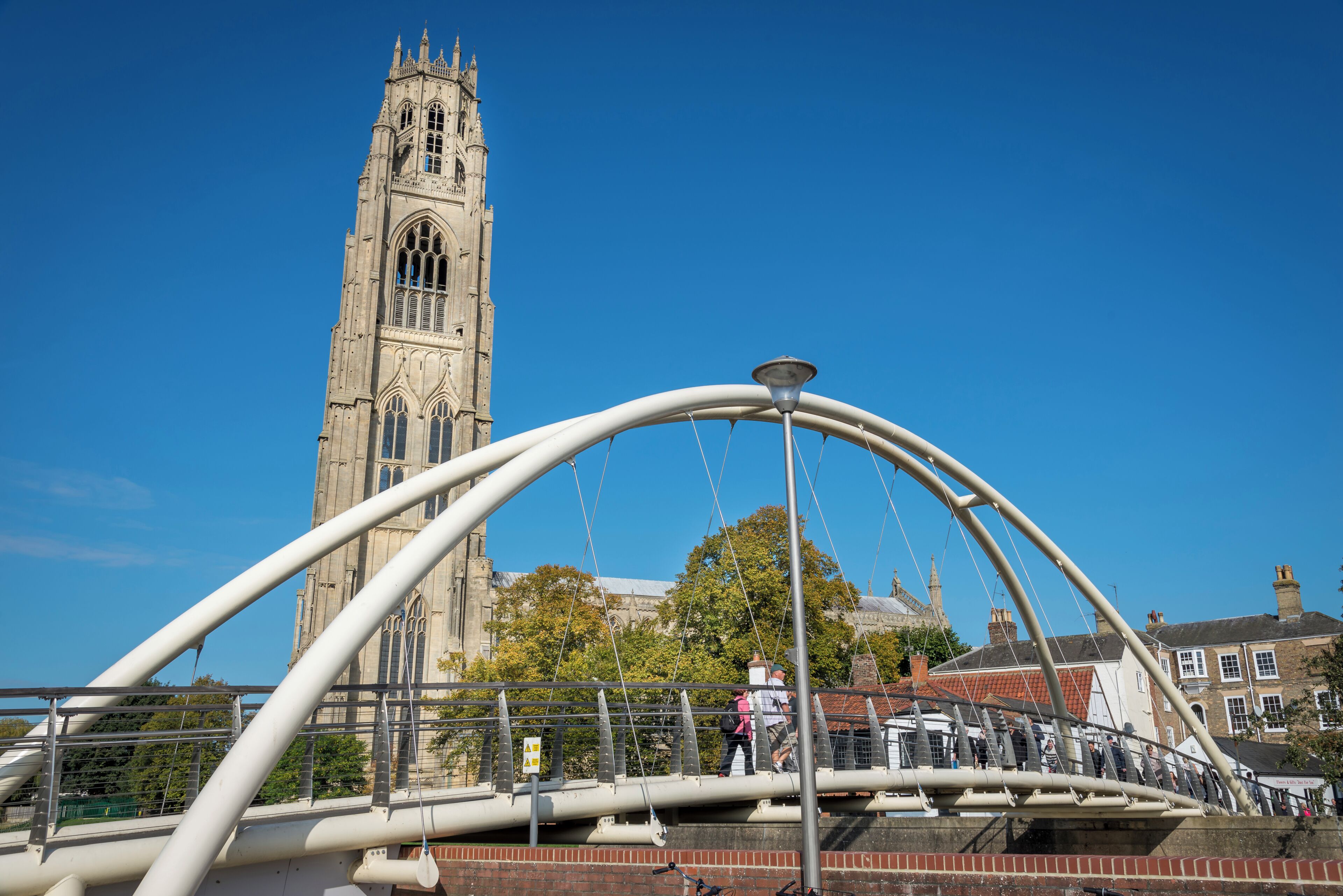 Un puente peatonal con estructura de arco metálico, una torre de iglesia histórica al fondo y un cielo azul despejado.