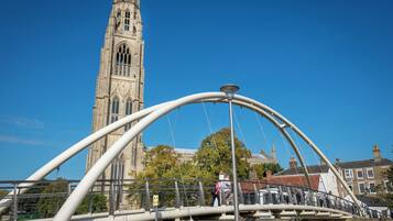 A pedestrian bridge with a metal arch structure, a historic church tower in the background, and a clear blue sky.