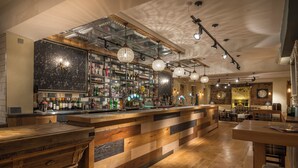 A bar area with a wooden counter, shelves with bottles, hanging lights, and a clock on the wall.