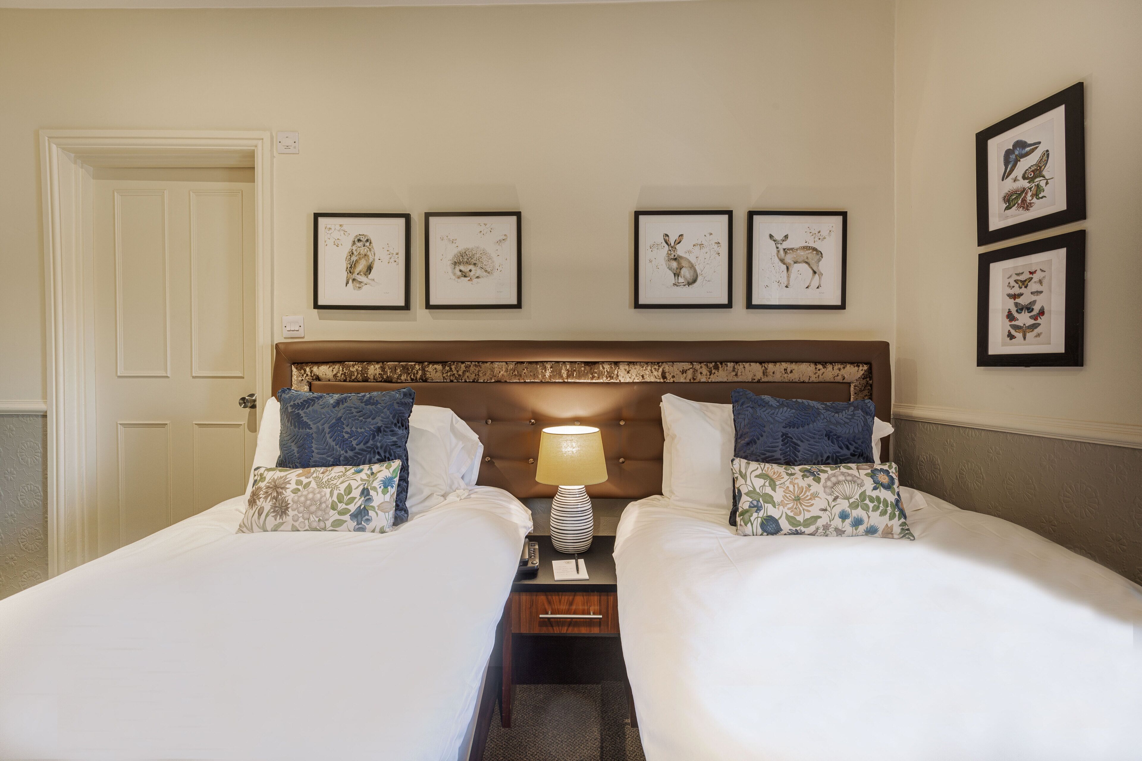 A hotel room with two beds, a nightstand with a lamp, and four framed animal illustrations on the wall.