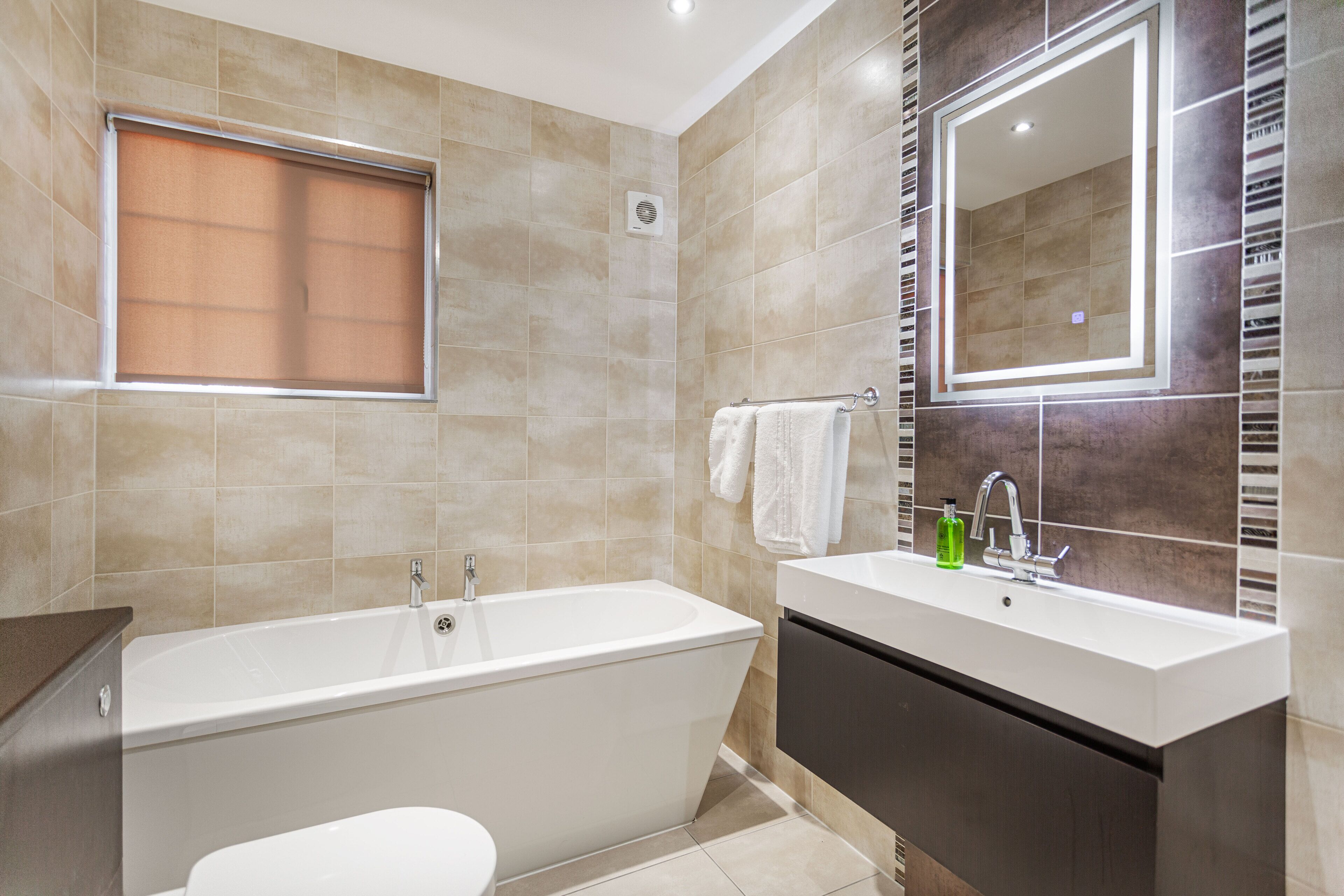 A modern bathroom with a bathtub, a sink, a mirror, and a window with a blind.