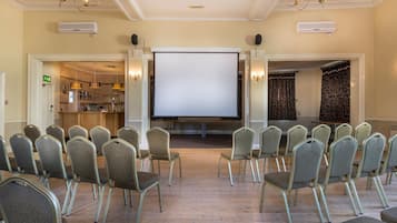 Un salón de conferencias con filas de sillas orientadas hacia una pantalla grande, una kitchenette y una cortina estampada.