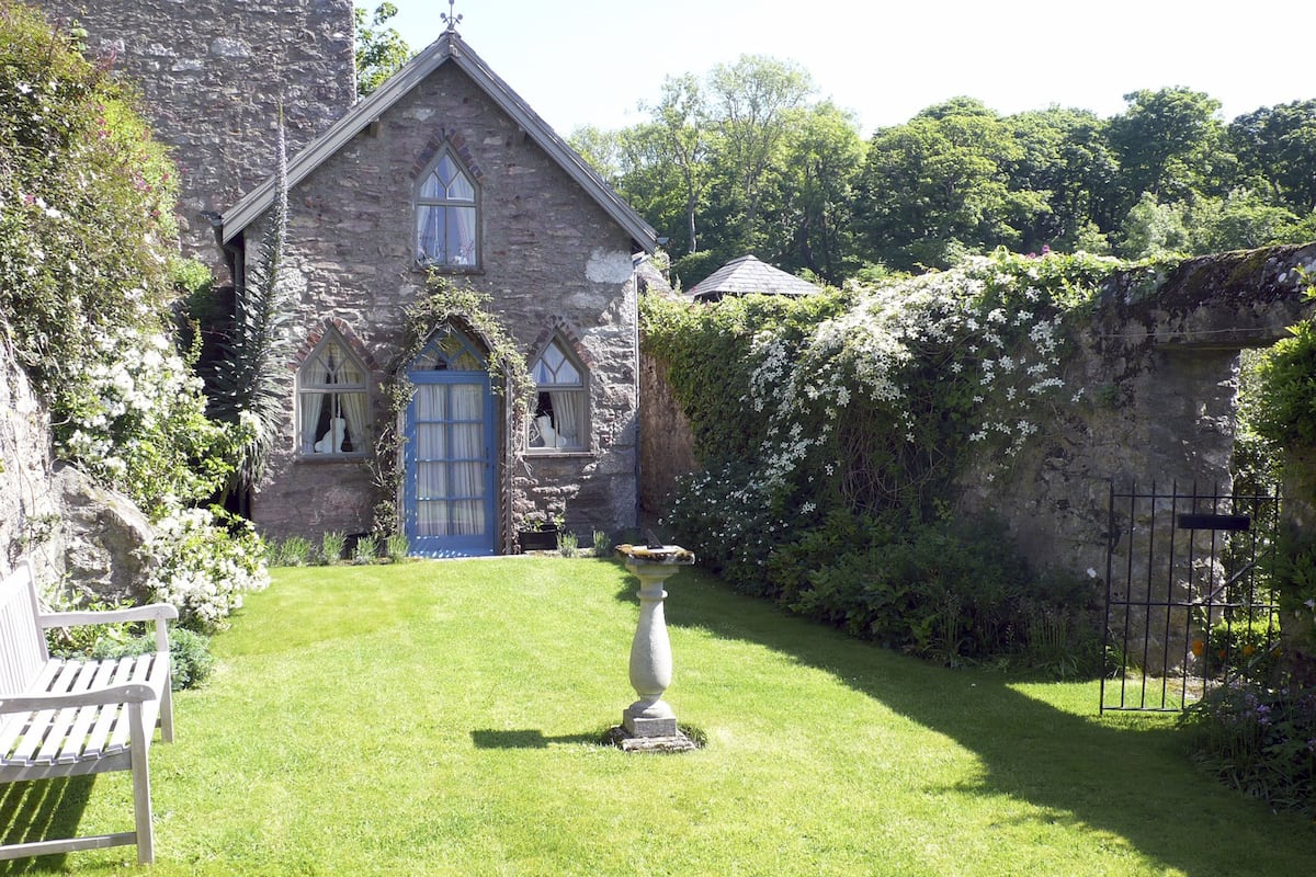 A stone building with a blue door and windows, surrounded by lush greenery and a small garden with a bench and a birdbath.