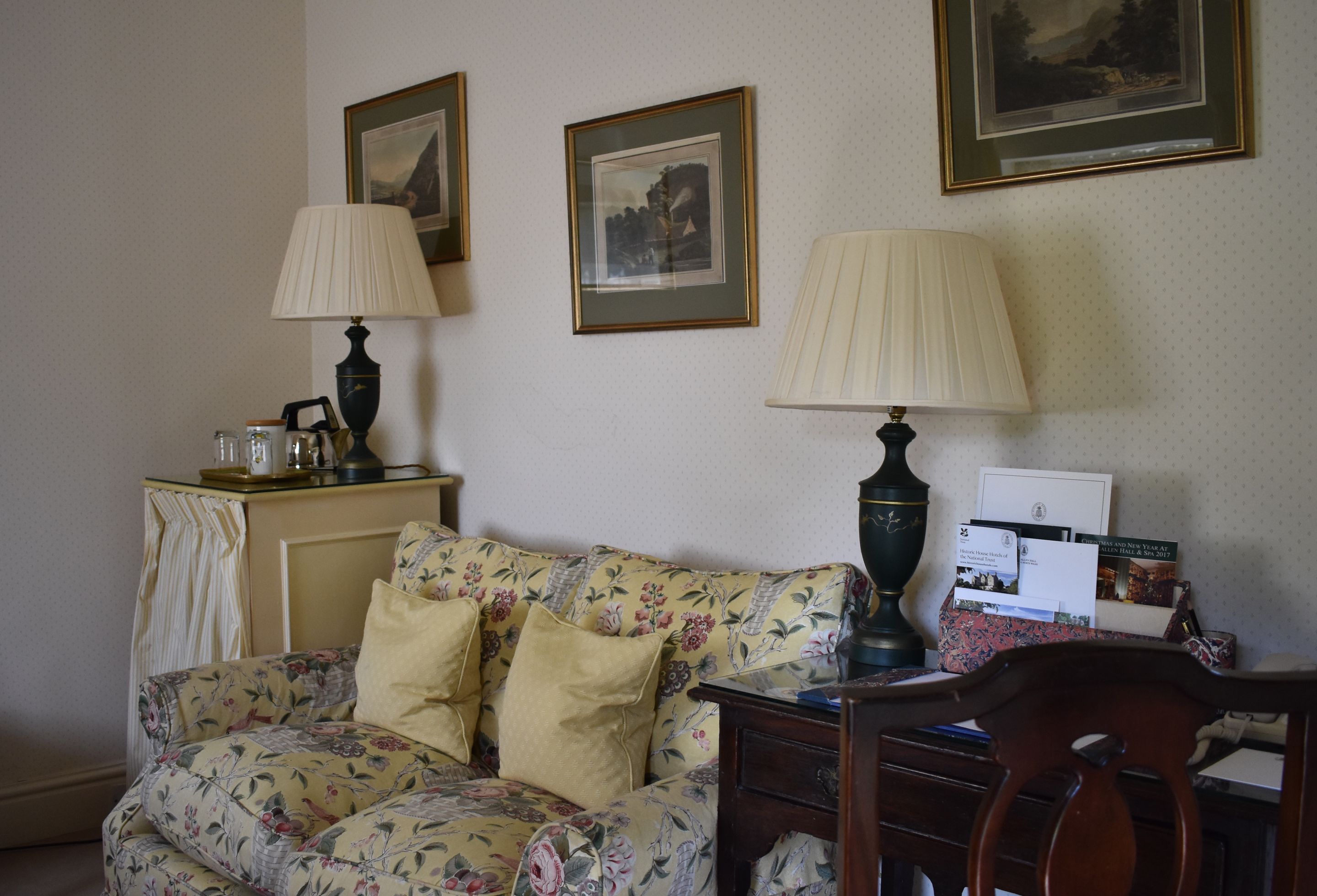 A room with a floral sofa, two lamps, a desk with papers, and framed pictures on the wall.