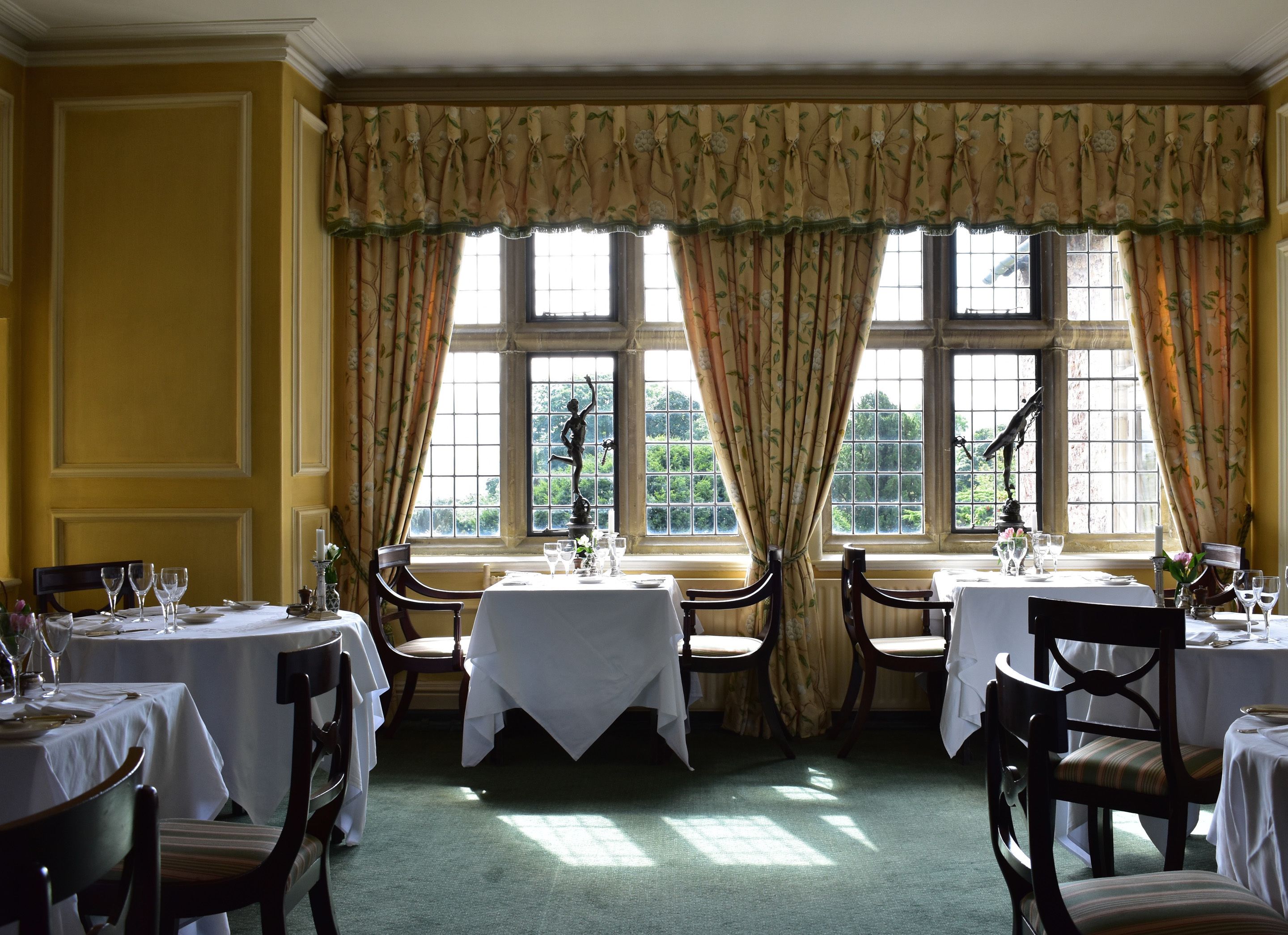 A dining area with multiple tables set for service, large windows with curtains, and a view of greenery outside.