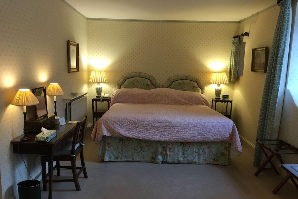 Un dormitorio con una cama doble, dos mesitas de noche con lámparas, un escritorio con silla y una ventana con cortinas.