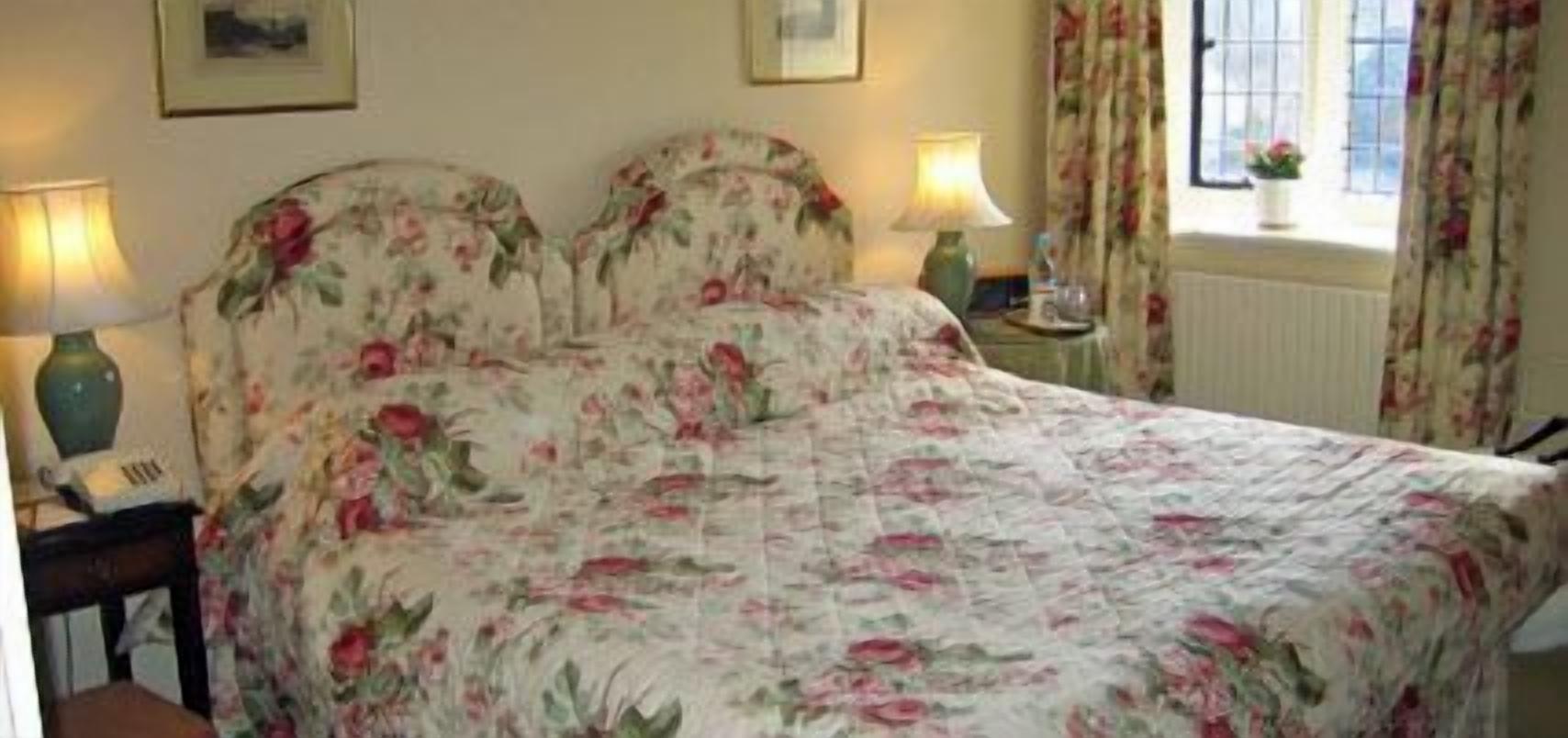 Een slaapkamer met een bloemenbedekking, twee bedlampjes, een nachtkastje en een raam met gordijnen.