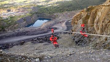 A person in red harness and helmet suspended on a cable, with a rocky terrain and a distant town visible.
