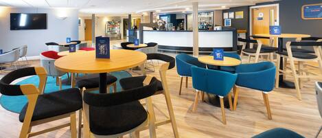 A modern restaurant with round tables and chairs, a bar area, and a television screen.