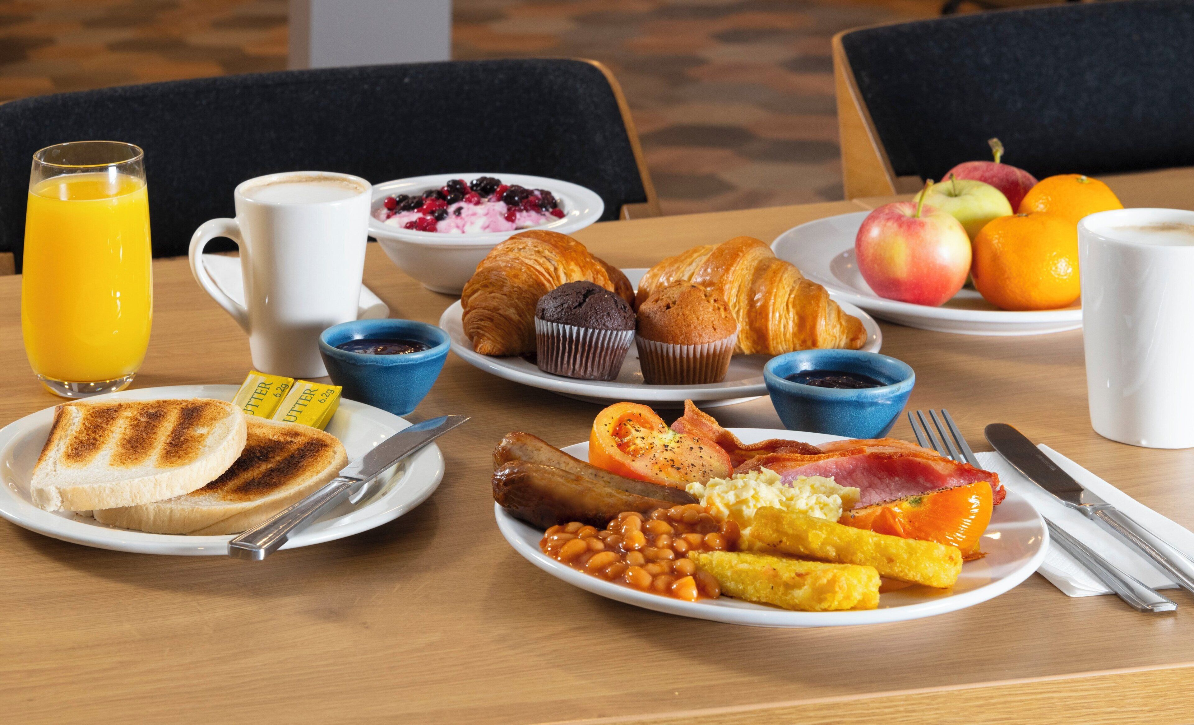 A breakfast table with plates of toast, muffins, beans, bacon, eggs, and fruit.