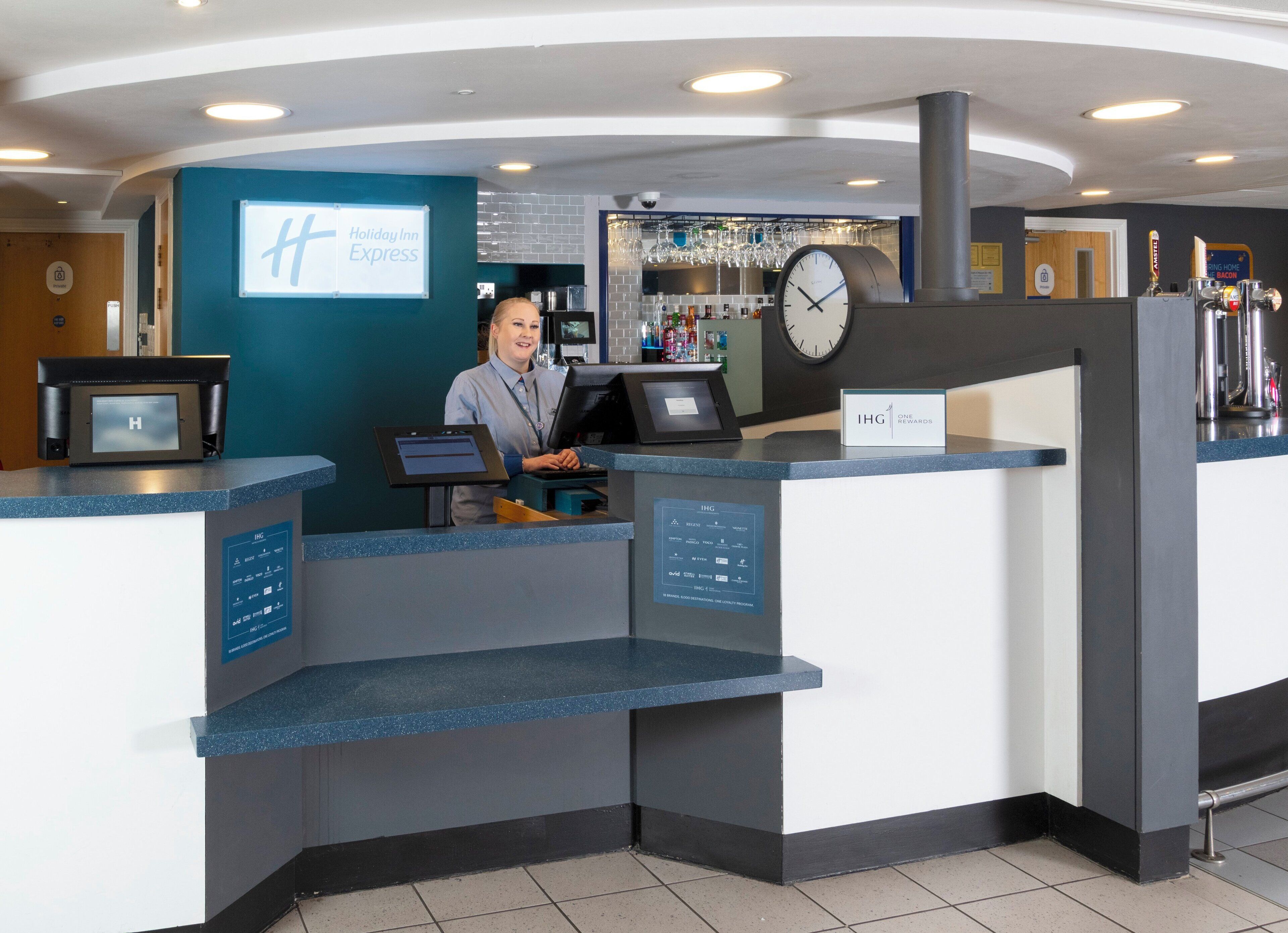 A hotel reception desk with a digital display, a clock, and a sign indicating a rewards program.