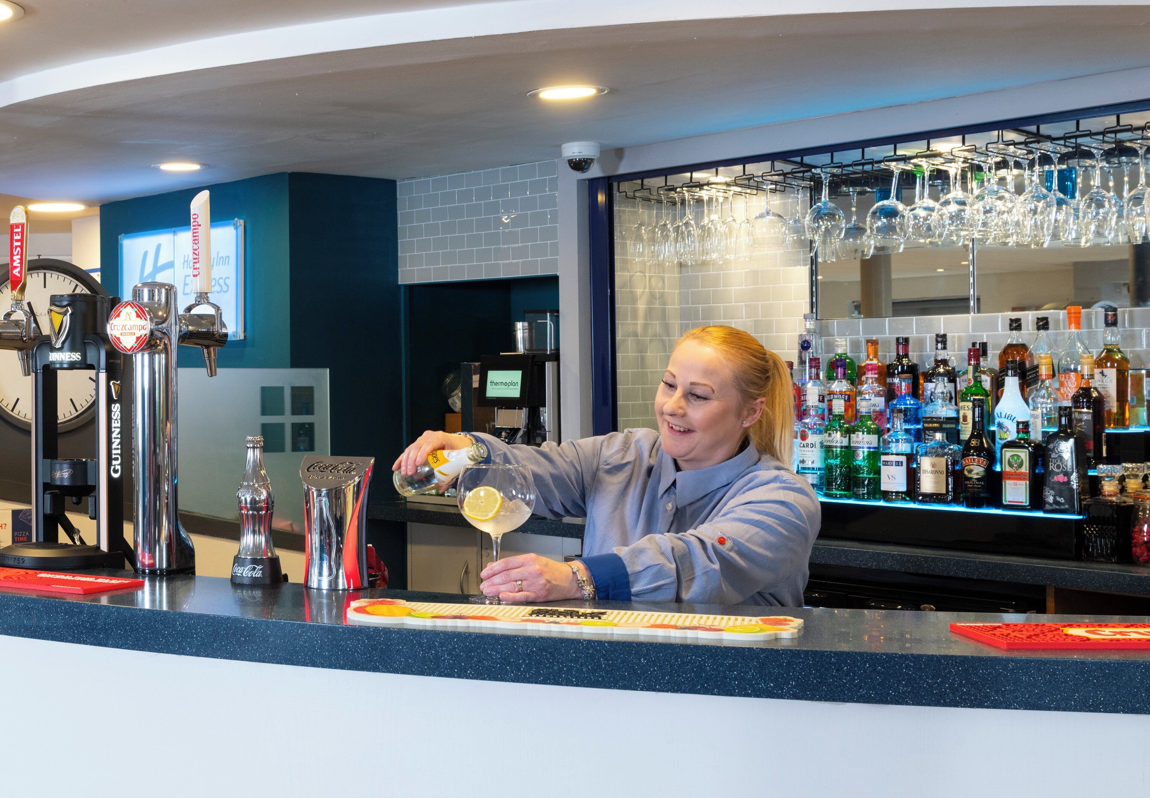 A bartender preparing a drink at a bar with a variety of liquor bottles and a clock on the wall.
