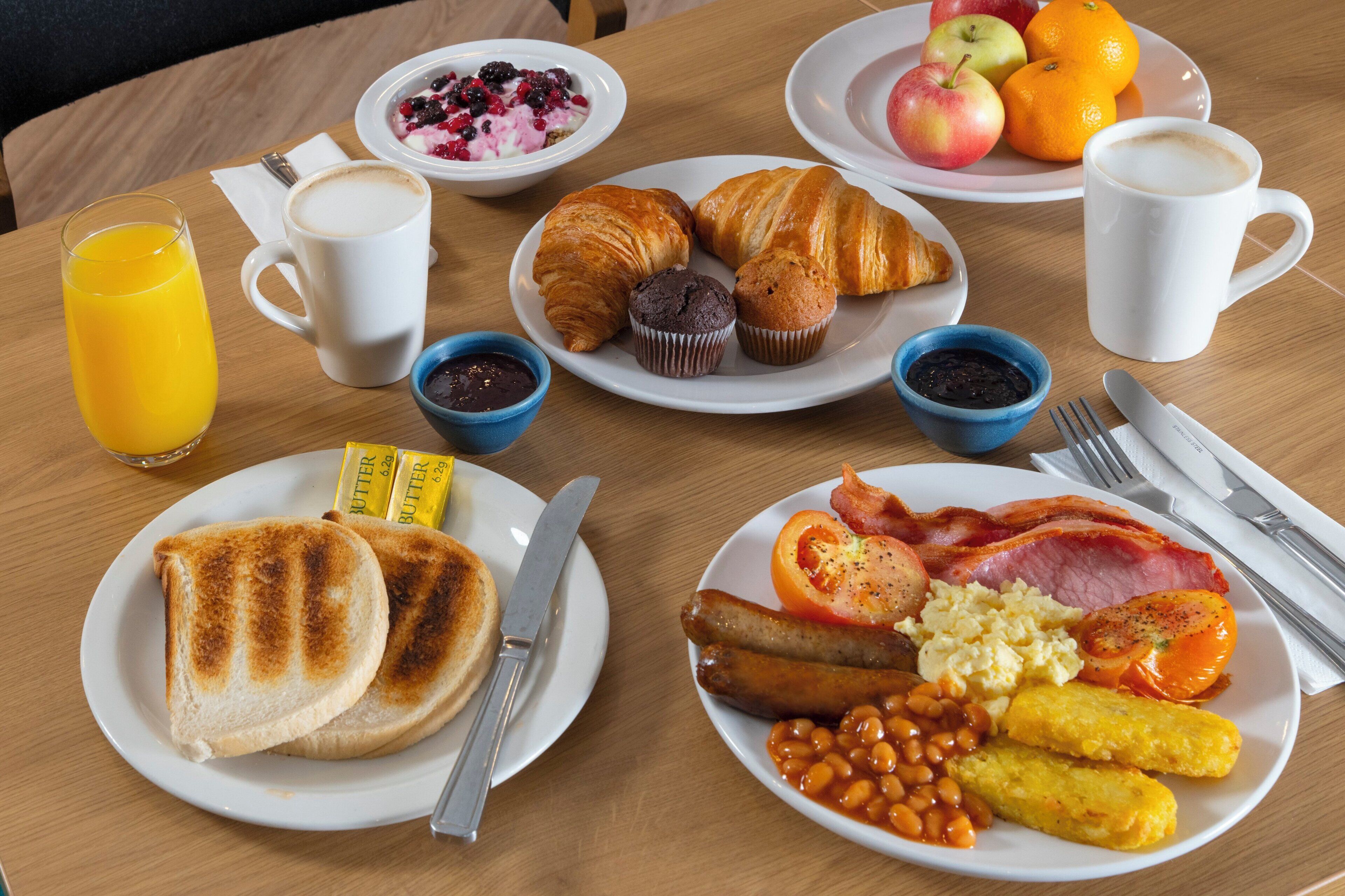 A breakfast table with plates of toast, eggs, bacon, beans, and baked beans. There are also croissants, muffins, and a bowl of fruit.