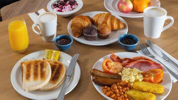 A breakfast table with plates of toast, eggs, bacon, beans, and baked beans. There are also croissants, muffins, and a bowl of fruit.