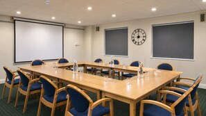 A conference room with a long table, blue chairs, a projector screen, a clock, and a water bottle on the table. - Holiday Inn Express Stafford by IHG (Stafford)
