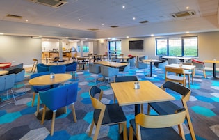 A modern indoor dining area with round tables and blue chairs, a kitchen in the background, and large windows providing natural light.