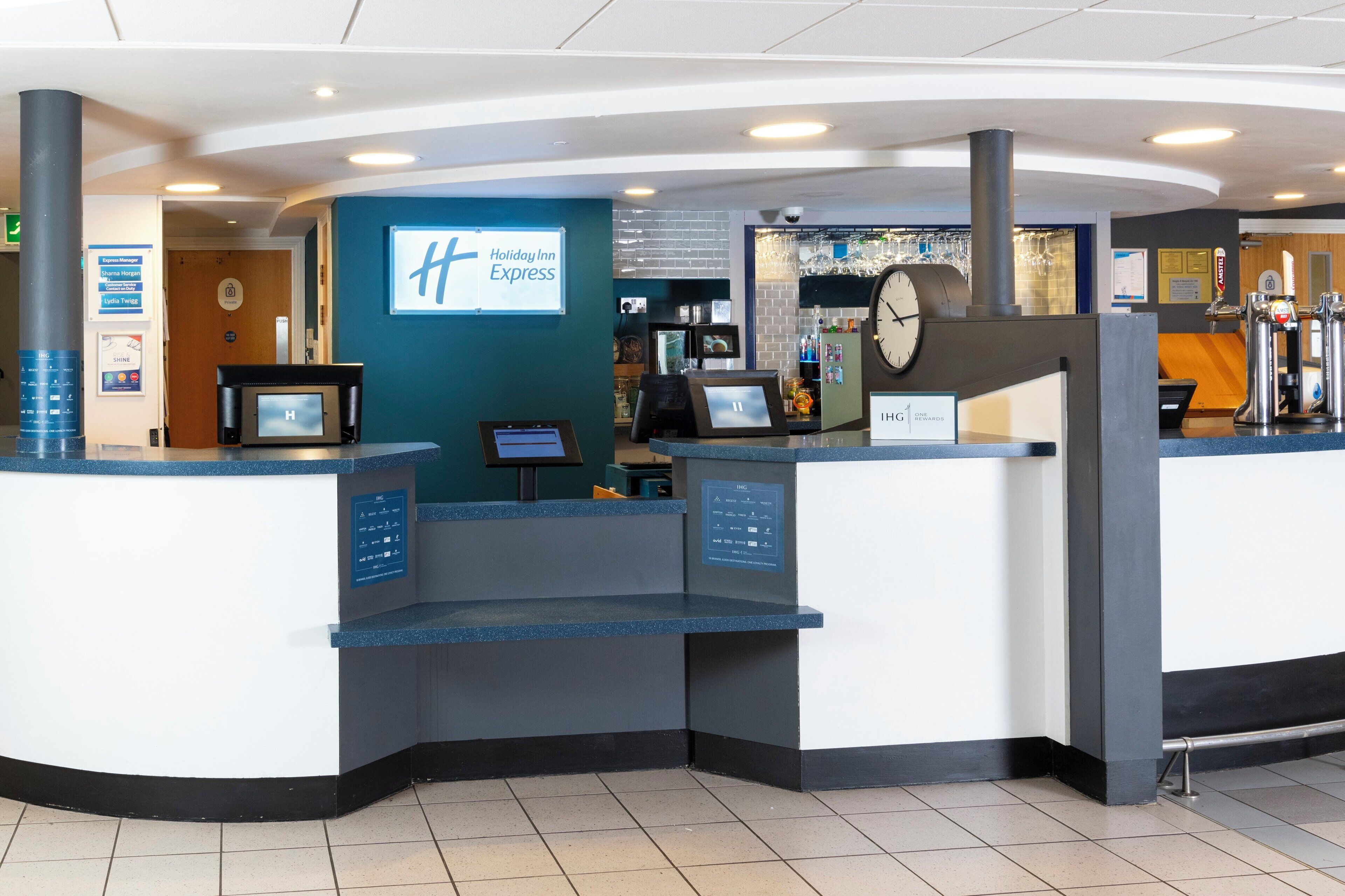 A Holiday Inn Express reception area with a digital display, a clock, and a bar area with glasses.
