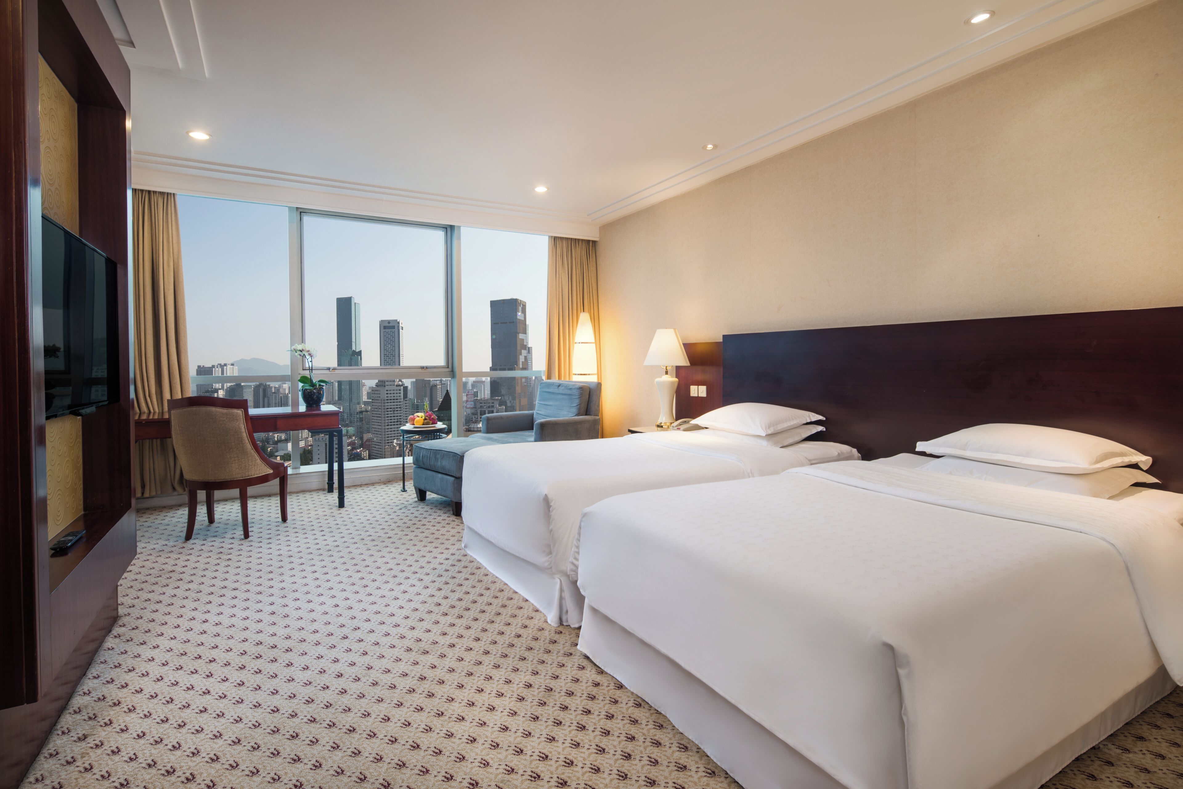 Habitación de hotel con dos camas, televisor, escritorio con silla y balcón con vista a la ciudad.