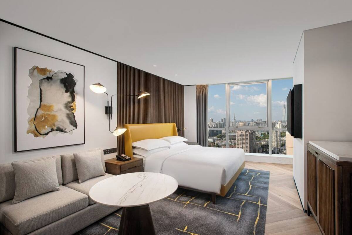 A modern hotel room with a bed, sofa, and a city view through large windows.