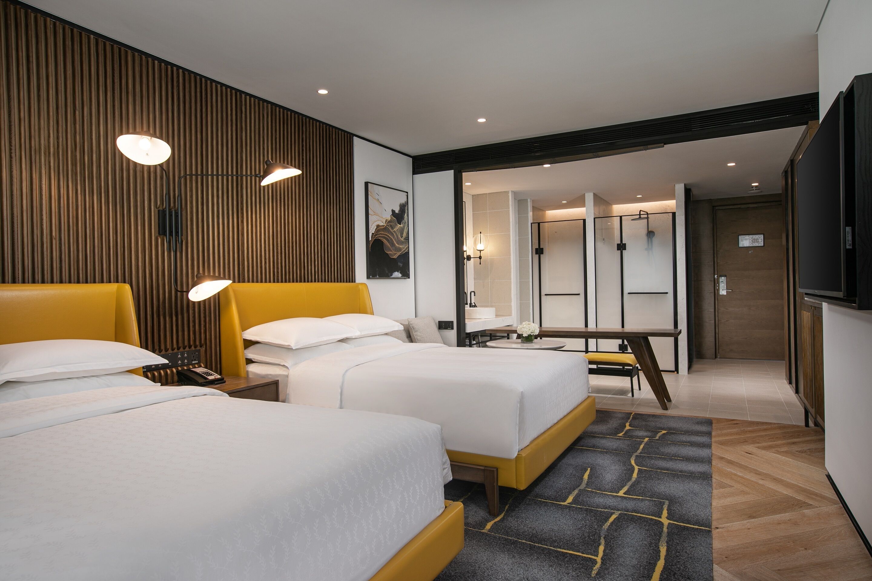 Ein Hotelzimmer mit zwei Betten, einer Wand mit Holzelementen, einem modernen Badezimmer und einem Flachbildfernseher.