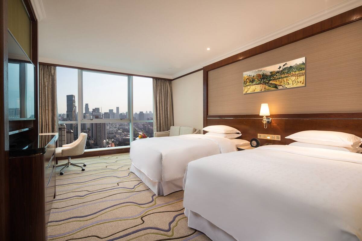 A hotel room with two beds, a desk, a chair, a TV, a large window with a city view, and a painting on the wall.