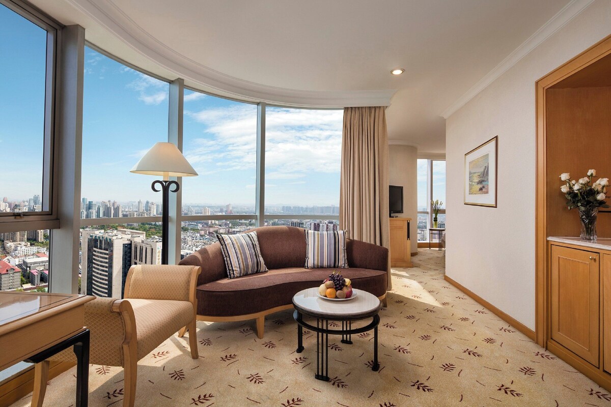 Habitación de hotel moderna con un sofá curvo, una mesita redonda con fruta, una lámpara y vistas a la ciudad.
