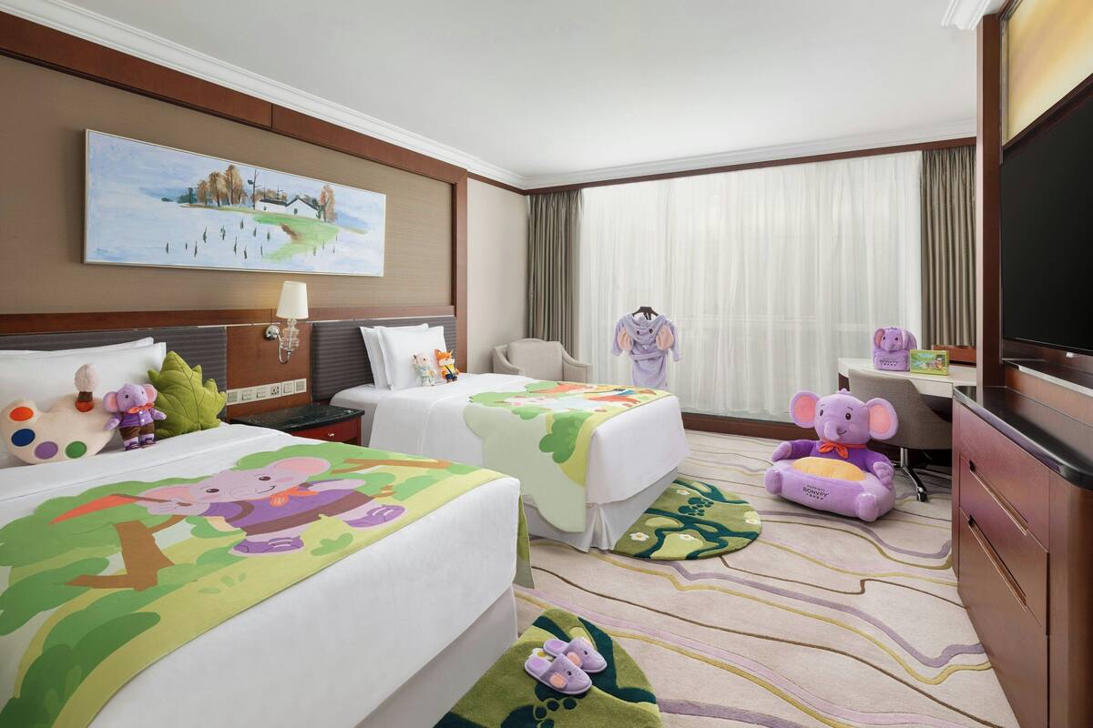 A hotel room with two beds, a television, a desk, and a plush toy elephant.