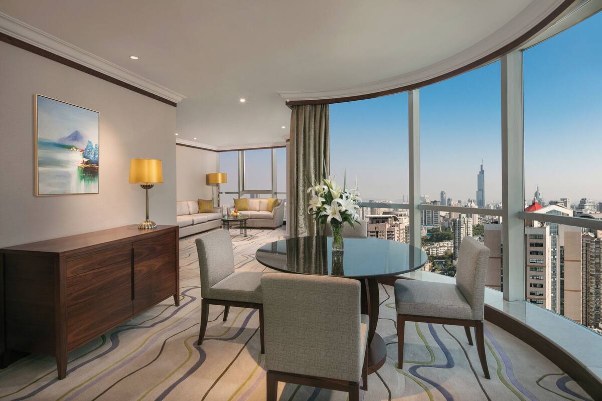A modern hotel room with a curved balcony, a dining table, two sofas, a coffee table, and a view of the city skyline.