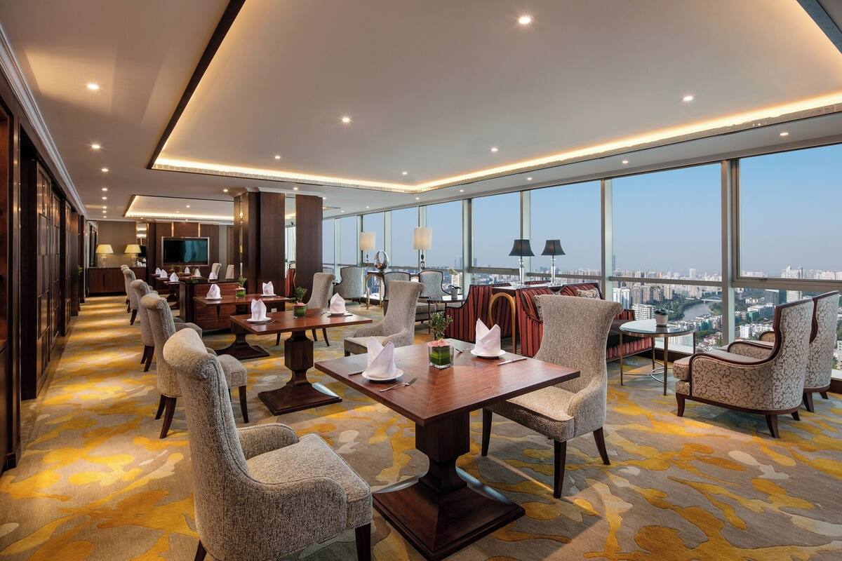 A modern hotel room with a large dining table, comfortable seating, a TV, and a city view.